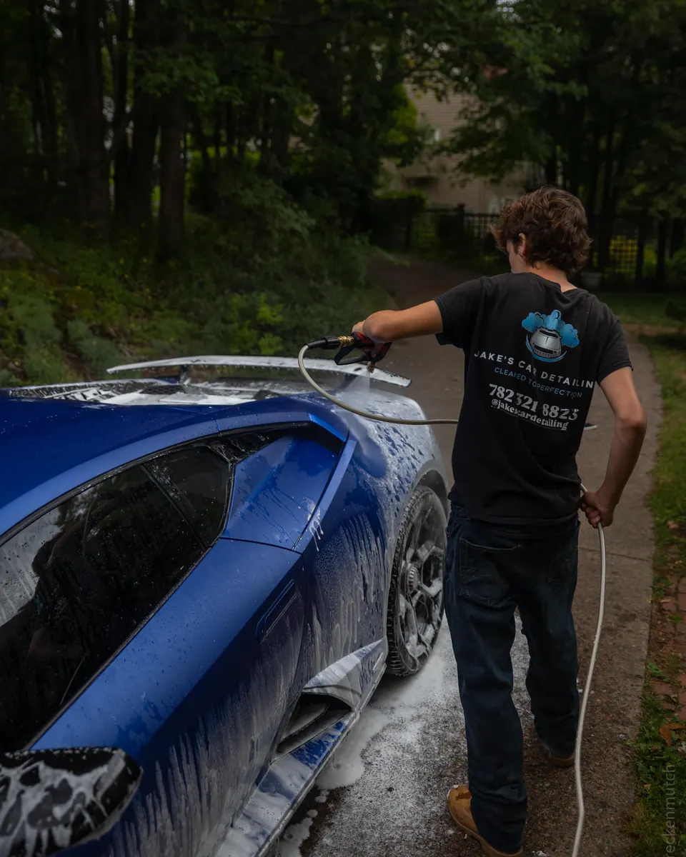 Jake rinsing snow foam off a blue Lamborghini Huracán on a tree-lined driveway